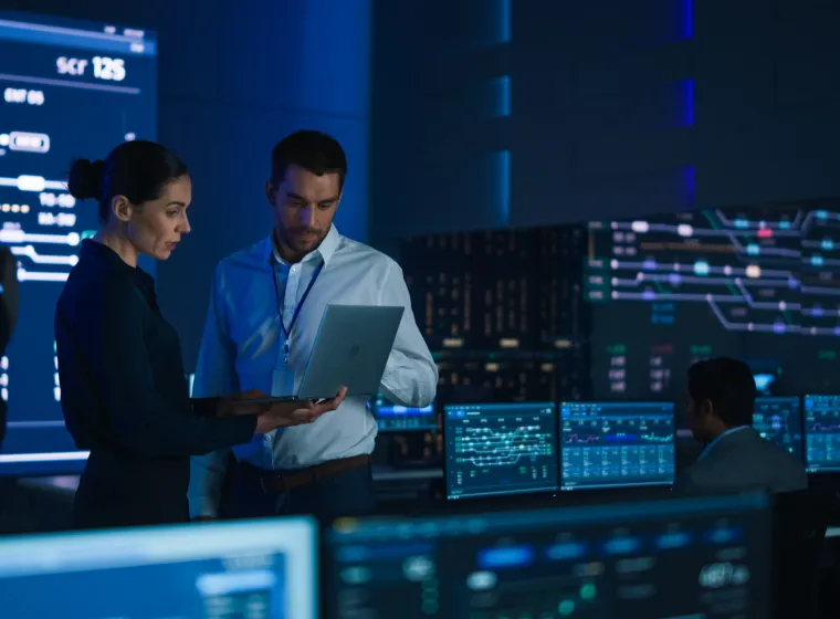 Project Leader and Chief Executive Discuss Data Shown on a Laptop. In Background Big Screen Showing Infographics of Infrastructure, Charts, System Analysis. Telecommunications Control Room