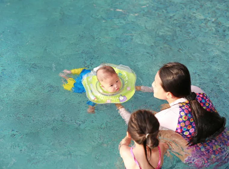  Mother and daughter training for infant baby boy in swimming suit floating in pool with safety by baby neck floats. 